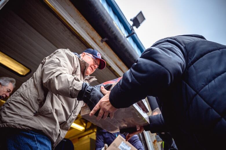 Two men, one in a beige jacket and a Montreal Canadiens cap and one in a blue jacket lift boxes into a truck at the loading bay of the food bank warehouse.