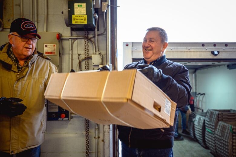 Two older men, one sporting a Montreal Canadiens cap and beige jacket and one wearing a blue jacket carrying a large cardboard box.