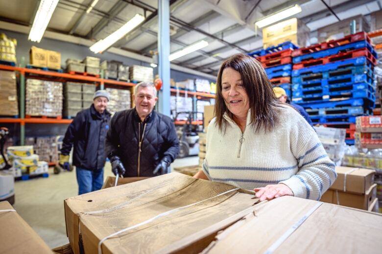 A woman with long brown hair prepares a cardboard box to be moved from the food bank warehouse. Two men in their seniors look on.