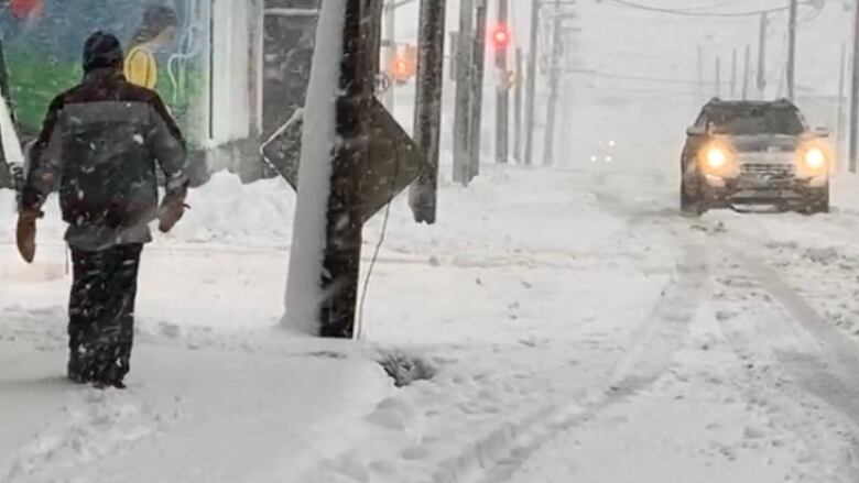 An SUV drives down a snowy road as someone walks through the snow on the sidewalk.