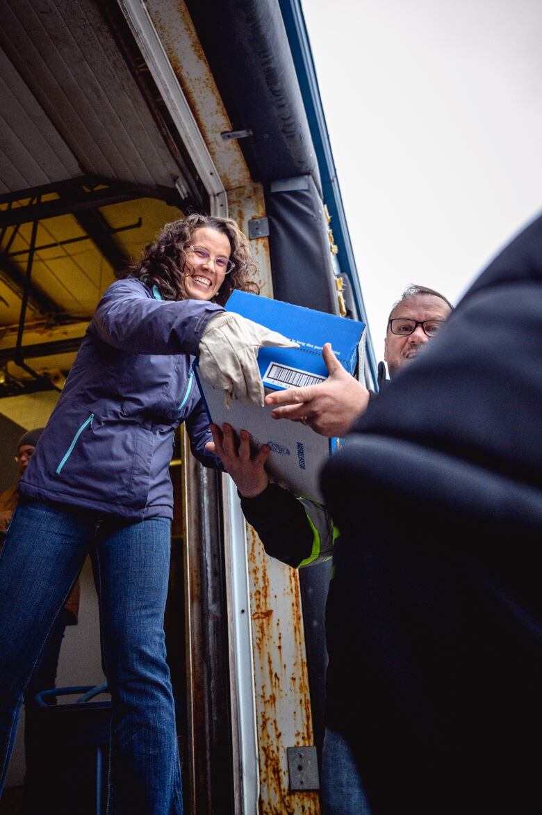 Woman with long brown curly hair and glasses smiles as she hands over a box of food to be loaded into a truck.