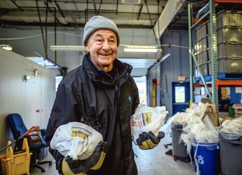 Older man smiling wearing a grey toque and blue jacket with two frozen turkeys in his hands.