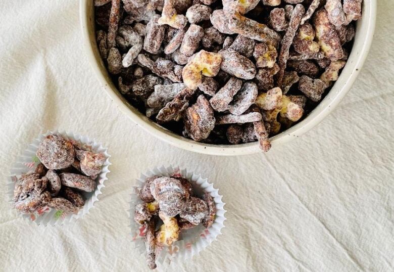 A bowl of chocolate-covered snack mix sits on a table next to two, small muffin liners filled with the same mix.