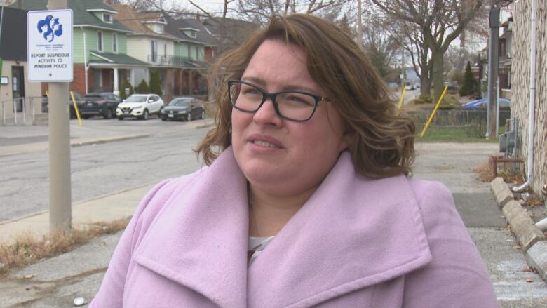 A woman in a pink coat and glasses stands on the sidewalk. She looks not happy. 