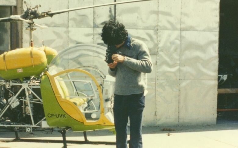 Young man is standing on a barrel with a vintage camera at the Delta Airfield. There's a helicopter and air hangar in the background. 