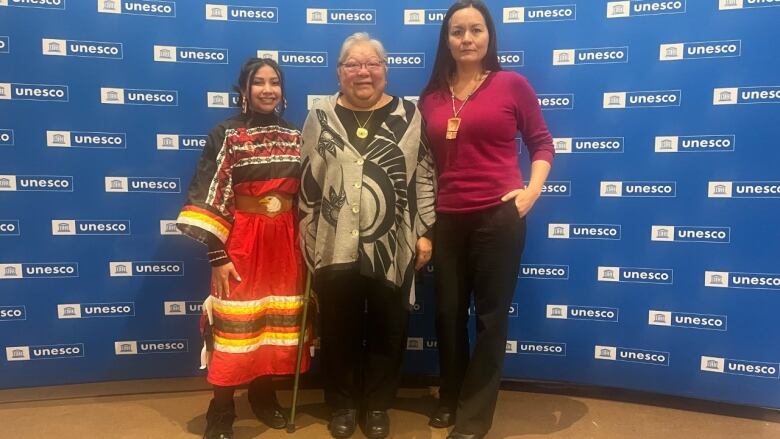 Three Indigenous women stand in front of a blue UNESCO back drop. The one on the left is wearing a red ribbon skirt. 