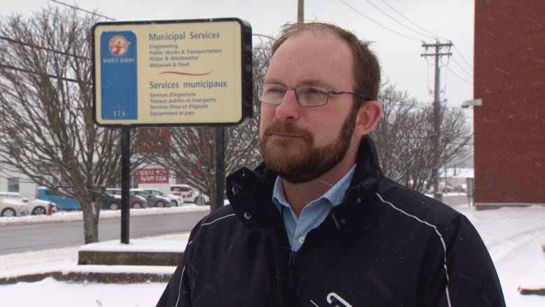 A man stands outside in the snow, we see him from shoulders up. He is wearing glasses, a blue button down, a black winter coat and has a beard. 