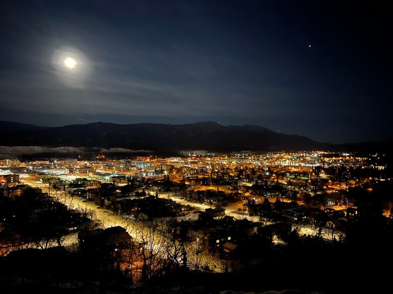 A view of a city at night, with the moon and mountains in the background.