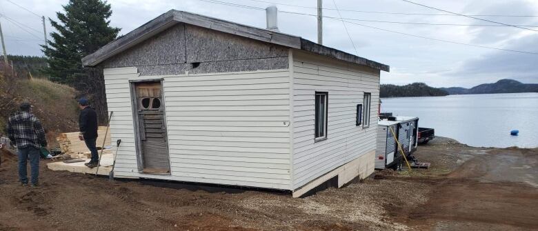 Two man stand next to a beige bungalow, which is under construction. The ocean is in the near distance. 