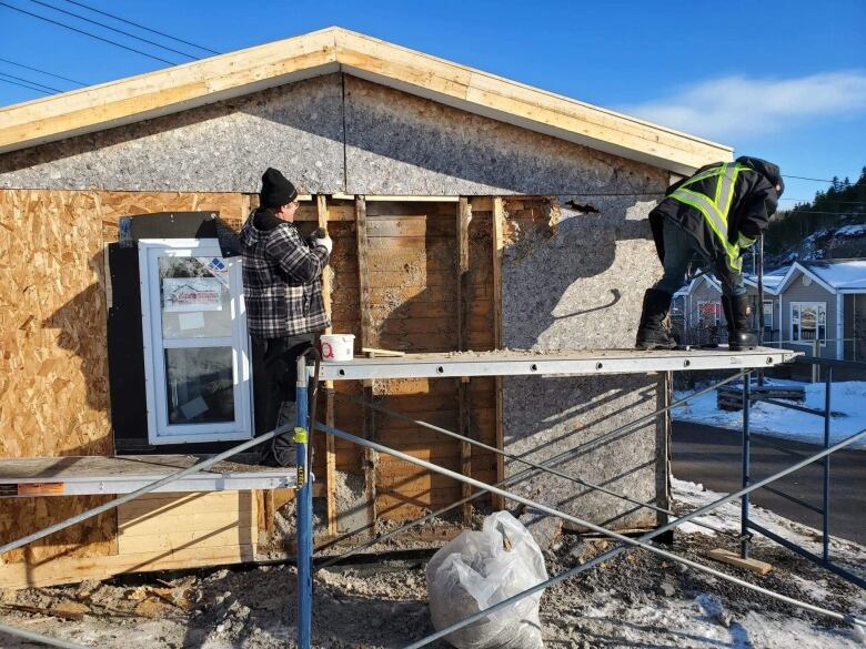 Two men stand on scaffolding, making repairs to a home. 