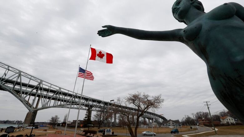 A statue with an outstretched arm is shown near a bridge connecting Canada and the United States.