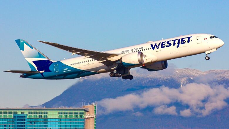 Blue and white WestJet plane is mid-air taking off from YVR airport. In the background is the YVR Fairmont hotel and snowcapped mountains covered in clouds. 