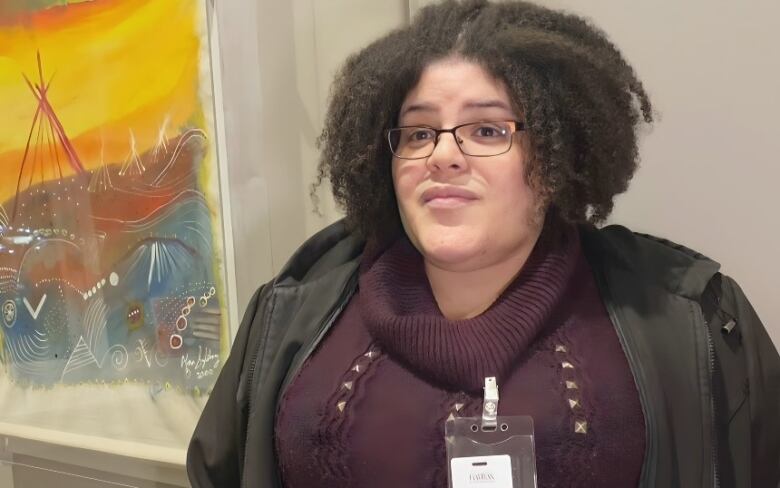 A Black woman in a jacket and purple sweater stands in front of a painting inside Halifax City Hall.