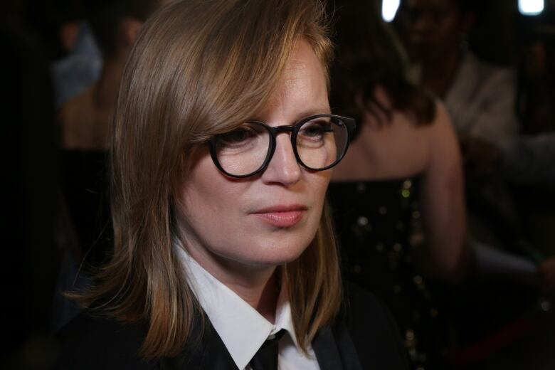 A close-up of a woman wearing thick black glasses, a dark blazer and tie over a white-collared shirt as she stands in a dimly lit room.