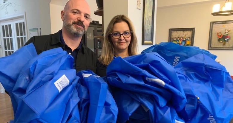 A man and a woman stand in their living room holding about 30 blue reusable Walmart bags they have collected from grocery delivery. 