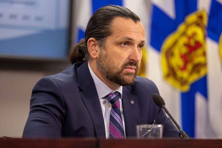 A man with a beard and ponytail sits at a conference table listening to questions.