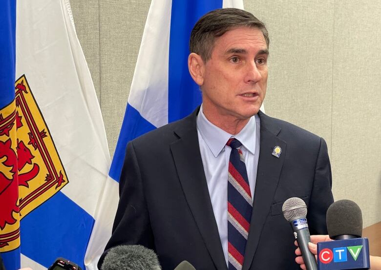 A man in a suit with dark hair stands at a microphone speaking to reporters.