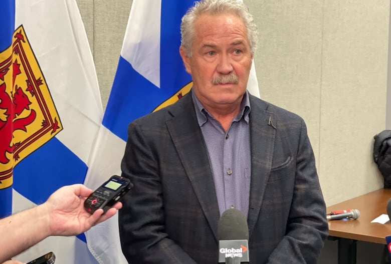 A man with white hair stands at a microphone speaking to reporters.