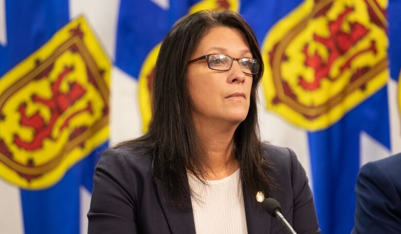 A woman with long black hair and glasses sits at a table and listens to questions.