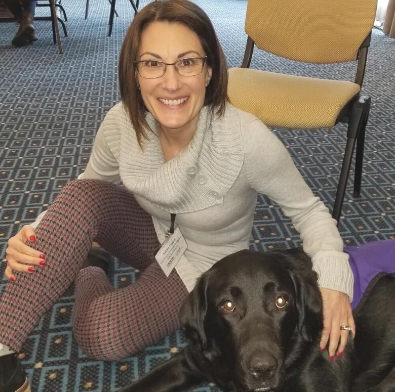 A photo of a smiling woman, seated on the floor beside a dog, The dog is wearing a blue jacket identifying it as a support animal.