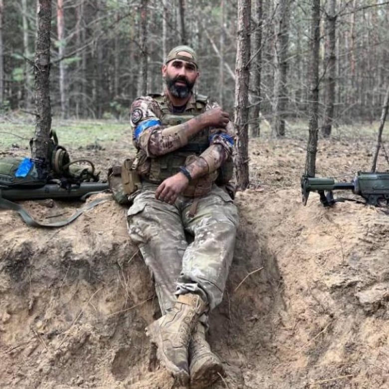 A man sits on the ground in the woods in camouflaged army gear and guns are laying beside him.