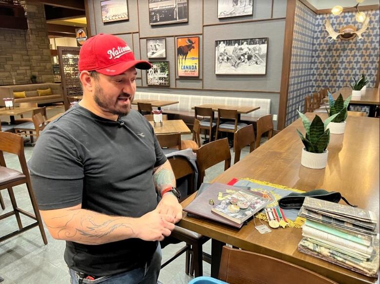 A man in a red ballcap stands near a bar counter. Medals, an army beret and old books are strewn across the countertop.