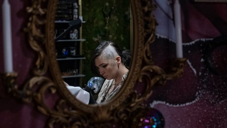 A reflection of a woman giving a tattoo is seen in a gold gilt frame mirror. Half of her head is shaved and it's tattooed.