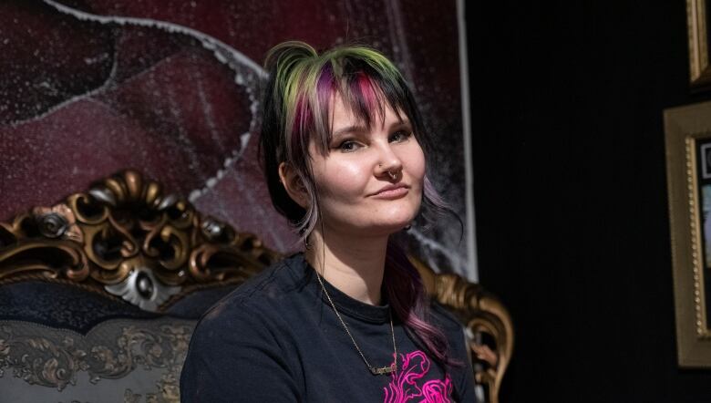 A young woman sits on an antique-style sofa. She's wearing a dark grey T-shirt with a hot pink design. She has green and pink hair, and a septum piercing.