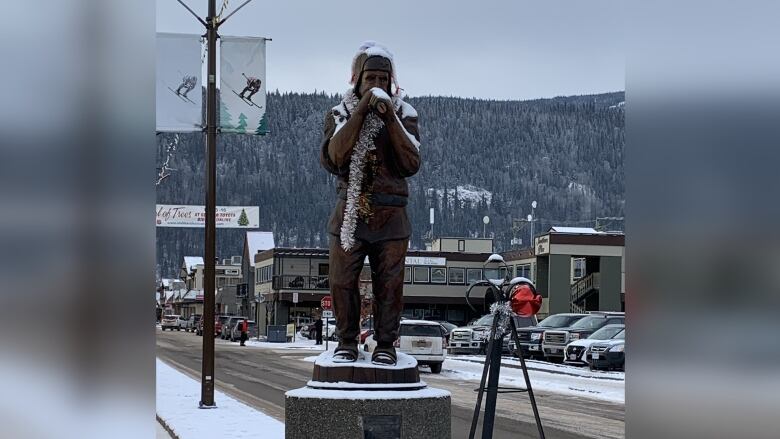 A statue in the shape of a man holding his hands around his mouth stands on a road covered with snow, with a tripod in front of it.