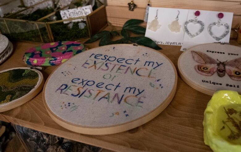 A needlepoint piece that says 'respect my existence or expect my resistance' sits in a wood frame on a table covered in other needlework and earrings.