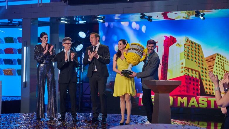 A woman in yellow and a man in grey suit are holding a trophy made of Lego bricks, while standing beside three other people on the stage of a studio.