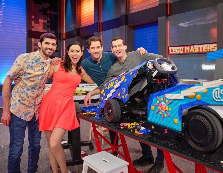 Three men and a woman stand next to a race car made of Lego bricks in a studio.