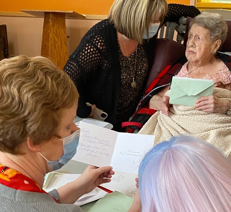 Two people sitting at a table, seen from behind, reading a card. Two people sit in front of them, seen from the front, and the one on the right holds an envelope.