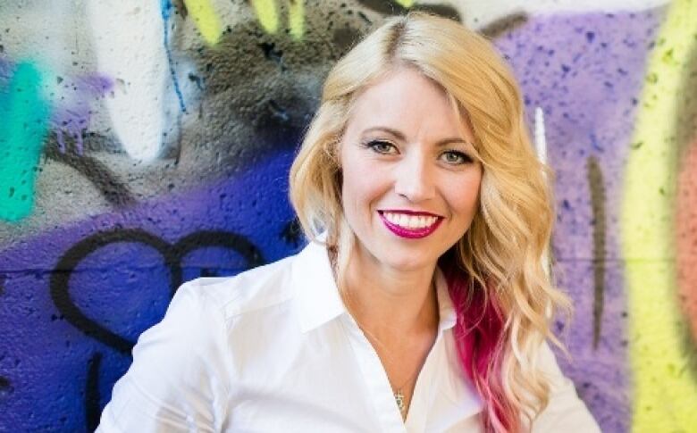 A smiling woman with blonde hair, streaked with pink, stands in front of a graffiti-covered wall.