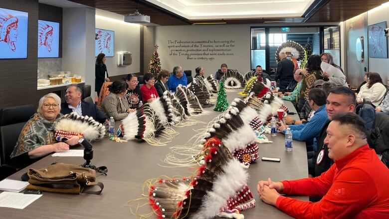 Several people sit at a long table in a conference room. Feathered headdresses sit on the table in front of many of the people.