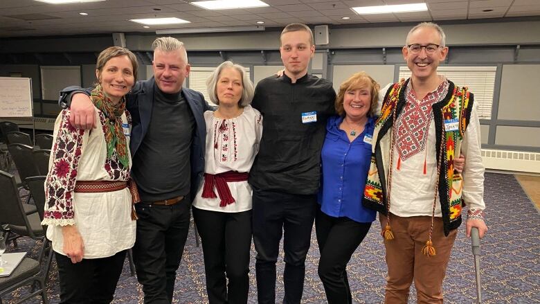 A group of smiling people, some dressed in Ukrainian clothing, stand in a line with their arms around each other.