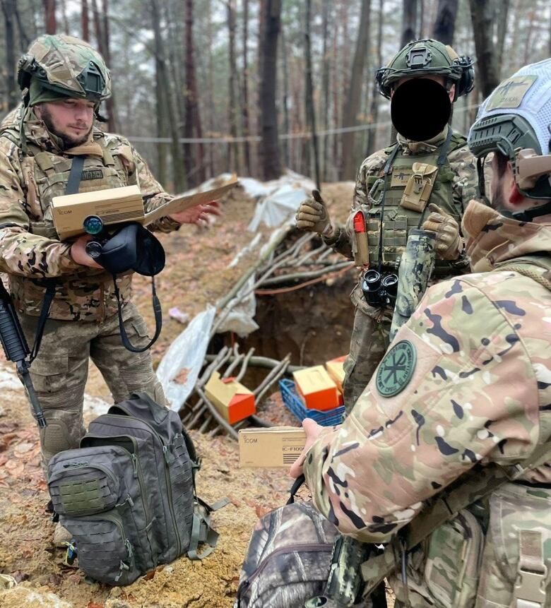 Three Ukrainian soldiers in camouflage gear stand in a circle in a forest, looking at some equipment.