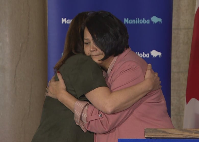 Two women embrace next to a podium. Behind them are a Canadian flag and a background with the Manitoba government's logo.