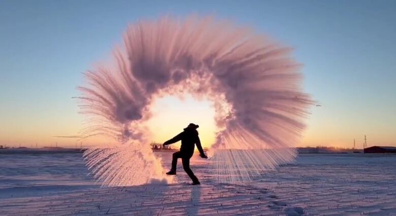 A man dances on a snow-swept, prairie landscape at dusk. He appears to be ringed by frozen mist as he dispenses water from a bottle all around him in icy cold weather.