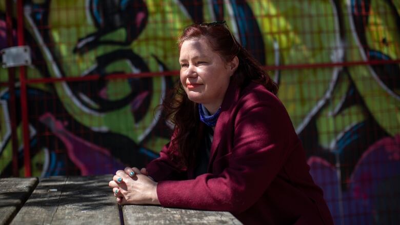 A woman with brown-red hair sits on a bench, with a graffiti wall behind her.