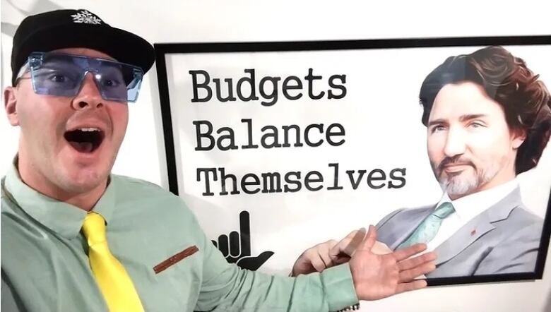 A man in a shirt and tie, wearing a ball cap, is smiling and gesturing towards a poster on a wall of Prime Minister Justine Trudeau with the caption Budgets Balance Themselves.