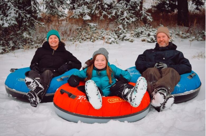 A family of three sits dressed in winter gear on tubes in the snow.