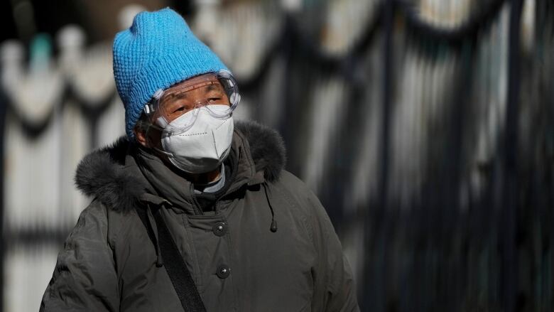 A man in a blue toque and black parka wears clear goggles and a face mask as he walks outside.