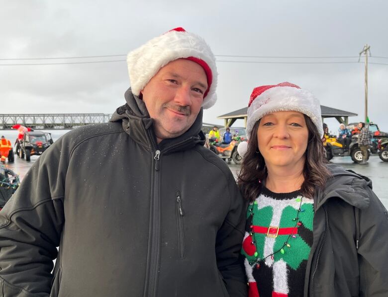 A man, left, and a woman, right, both wearing Santa hats, are smiling for the camera. In the background, there are people and ATVs, which are decorated with lights and Christmas inflatables.