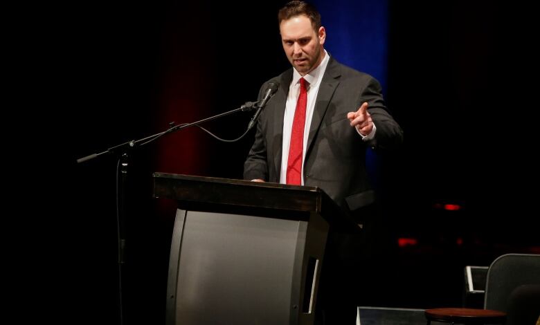 A man in a dark suit and a red tie points as he speaks at a podium.