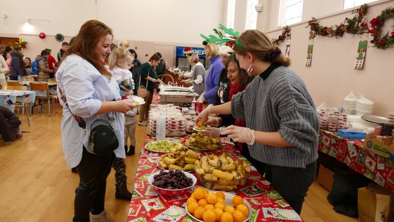 A woman in a light blue shirt stands at a buffet style table. Another woman, in a grey sweater, hands her food.