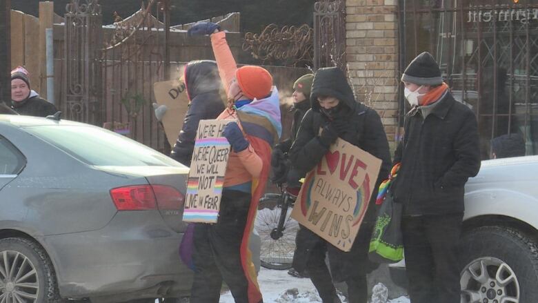 Standing in front of parked cars, people in winter attire hold signs reading 