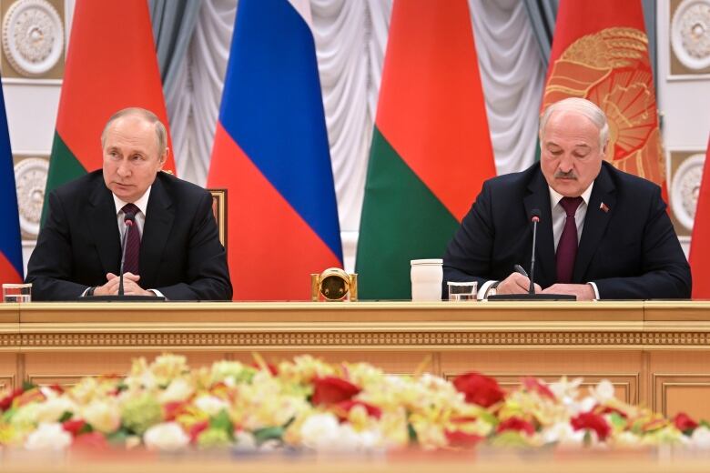 Two men sit behind a wooden table, in front of a row of blue, red, green and gold flags. One man is looking ahead while the other is looking down and appears to be writing.