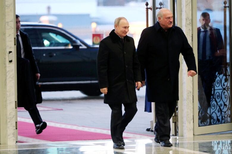 Two men in black coats and black pants walk into a building which appears to have marble floors and walls. The shorter man is smiling. Behind them is a red carpet, and two men who appear to be bodyguards, who are dressed in black suits and black ties.