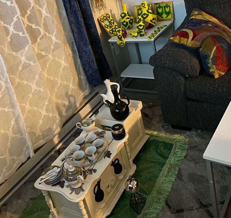 A corner of a room is pictured with a green rug, a dark couch, a straw decorative wall ornament, and a white coffee table with white cups and a jug.